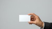 Close-up of male hand in dark business suit holding blank white card against neutral gray background for corporate branding and professional networking purposes.