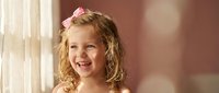 Young blonde girl with curly hair and pink bow smiling joyfully by window with soft natural lighting creating warm atmosphere for family portraits.