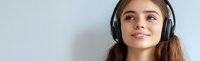 Young woman with long brown hair wearing black wireless headphones smiling and looking up while listening to music against light gray background.