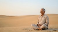 Senior man with gray hair and light skin tone, wearing casual beige clothing, meditating cross-legged on mat in vast desert landscape during golden hour. Concept for mindfulness, wellness, and spiritual practice.
