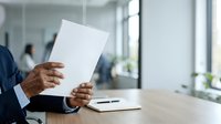 Mature male professional in dark business suit holding blank white document at wooden desk in modern office environment with colleagues and windows in background.