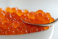 Fresh red caviar in white ceramic bowl with silver spoon, close up view showing translucent orange spheres with natural light highlighting their glossy texture.