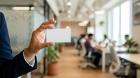 Businessman holding blank white business card in modern office environment with colleagues working in background, suitable for corporate branding and professional networking concepts.