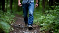 Person walking alone on forest trail wearing blue jeans and hiking boots, low angle view focused on legs. Green foliage surrounds narrow dirt path in woods.