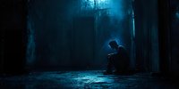 Silhouette of lonely person sitting in dark corridor with dramatic blue moonlight streaming through window, creating moody atmospheric scene.