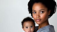 Young African American mother in gray scrubs holding baby, both looking directly at camera against light background. Professional healthcare worker portrait.