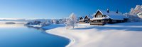 Cozy wooden cabin covered in snow beside frozen lake with snow-capped mountains under blue winter sky. Perfect for holiday cards and winter vacation concepts.