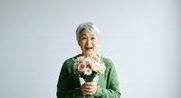 Senior woman with gray hair and Asian ethnicity, wearing a green cardigan, holding a bouquet of pink and white carnations against a white background. Suitable for greeting cards, Mother's Day promotions, senior lifestyle content, and wellness campaigns.