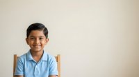 Young boy with dark hair and South Asian ethnicity wearing light blue collared shirt, seated on wooden chair against neutral beige background. Suitable for educational materials, school promotions, diversity campaigns.