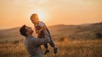 Father lifting young son in golden sunset field creating joyful family bonding moment during outdoor playtime in countryside landscape.