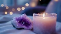 Glowing candle in glass holder next to purple amethyst crystal cluster on soft fabric with bokeh lights creating magical spiritual atmosphere for meditation and wellness.