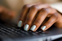 African American woman's hands with light blue manicure typing on black computer keyboard, shallow depth of field focus on well-groomed nails at work.