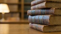 Stack of aged leather-bound books with decorative spines on wooden table, warm library setting with lamp and shelves blurred in background.