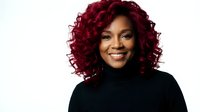 Professional African American woman with vibrant burgundy curly hair wearing black turtleneck sweater smiling confidently against white background.