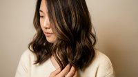 Young Asian woman with highlighted wavy hair touching her locks in cream sweater against neutral background for hair care and beauty concepts.