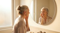 Middle-aged Caucasian woman with blonde hair in bathrobe looking at herself in round bathroom mirror, touching face during morning skincare routine.