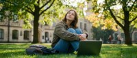Young woman student sitting on grass with laptop computer outdoors on university campus during sunny day, smiling and looking up thoughtfully.
