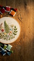 Botanical embroidery hoop with colorful floral and fern stitching on linen, surrounded by thread skeins and needles on rustic wood.
