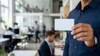 Man in navy shirt holding blank white business card in modern office environment with colleagues working in background.