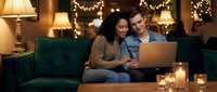 Young diverse couple using laptop together on cozy sofa with warm lighting and candles creating romantic atmosphere for online shopping or entertainment.