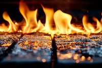 Bright orange flames dance across burning embers and ash, creating dramatic contrast against dark background. Close-up macro photography captures fire's natural intensity.