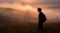 Young man silhouette standing in mountain meadow at golden sunrise with misty valleys and dramatic cloudy sky creating peaceful contemplative mood.