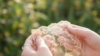 Delicate lace fabric held gently in cupped hands against soft natural bokeh background, showcasing intricate floral patterns and feminine textile craftsmanship.