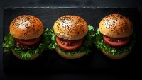 Three fresh hamburgers with sesame buns, juicy tomato slices, crisp lettuce on dark slate background, dramatic lighting creates appetizing mood for menu or food advertising.