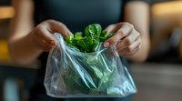 Fresh green spinach leaves being placed in plastic produce bag at grocery store, shallow depth of field with focus on transparent bag and vibrant greens.