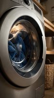 Modern stainless steel front-load washing machine with blue towels inside, set in a cozy laundry room with wooden shelves and wicker basket.