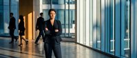 Professional businesswoman with crossed arms standing confidently in modern office corridor with glass walls and blurred colleagues in background.