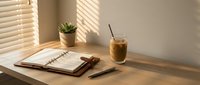 Wooden desk workspace with open notebook, pen, succulent plant in pot, and iced coffee drink by sunlit window with blinds casting shadows.