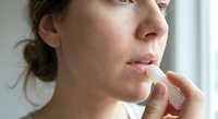 Young woman applying lip balm stick to lips for skincare routine and moisture protection in natural daylight near window.