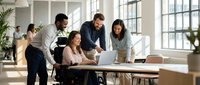 Diverse business team collaborating on laptop in modern office workspace with large windows and natural light for corporate presentations.