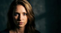 Intense portrait of young Caucasian woman with piercing blue eyes and flowing brown hair against dark moody background for dramatic storytelling concepts.