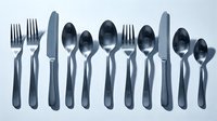 Stainless steel cutlery set with forks, knives, and spoons arranged neatly on a light background, casting soft shadows.