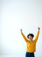 Joyful young woman with dark curly hair, medium brown skin tone, wearing mustard yellow sweater and blue jeans, arms raised in celebration against white background. Perfect for success, achievement, happiness and motivation concepts.