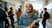 Young Muslim female student wearing hijab and denim jacket smiling while holding books in busy school hallway with diverse classmates in background.