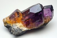 Raw amethyst and citrine crystal cluster formation on matrix stone showing natural color gradient from deep purple to golden amber, mineral specimen on light background.