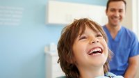 Happy young boy laughing during dental checkup with smiling male dentist in blue scrubs at modern clinic office for pediatric oral health care.