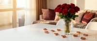 Romantic bedroom setup with red roses in vase and heart-shaped chocolates on white bed for Valentine's Day celebration and intimate moments.