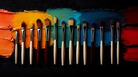 Professional makeup brushes arranged in row on colorful paint strokes background showing different shades of eyeshadow swatches, creative cosmetic flatlay composition.
