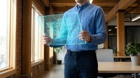 Male professional in blue shirt holding glowing digital tablet displaying holographic data visualization in modern office workspace with wooden beams and natural light.
