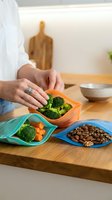 Adult hand holding colorful silicone food storage pouches filled with fresh broccoli, carrots, and legumes on wooden kitchen counter. Sustainable meal prep and eco-friendly kitchen storage solution.