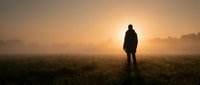 Person standing in misty field during golden sunrise with warm atmospheric lighting and peaceful countryside landscape.