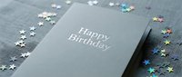 Dark elegant birthday card with silver foil lettering surrounded by colorful holographic star confetti on a gray textured surface.