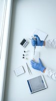 Scientist in white lab coat and blue nitrile gloves conducting medical research with test tubes, pipette, and digital equipment on white surface for laboratory analysis.