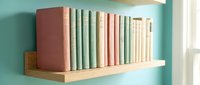 Pastel colored vintage books arranged on a wooden shelf against a teal wall, creating a cozy and stylish home library decor scene.