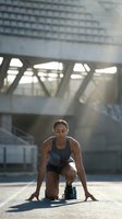 Athletic Hispanic woman in starting position ready to sprint on urban track with modern stadium architecture in background during golden hour training session.