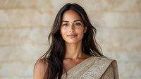 Young South Asian woman wearing traditional beige sari with embroidered pattern smiling warmly at camera against neutral wall background. Natural makeup and long dark hair.